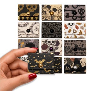 May include: A collection of twelve small, rectangular paper envelopes with snap closures, featuring various dark and gothic-inspired patterns including skulls, bats, keys, autumn leaves, and floral motifs in black, gold, white, and muted earth tones.