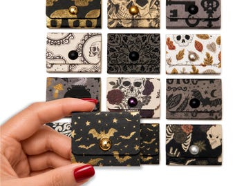 May include: A collection of twelve small, rectangular paper envelopes with snap closures, featuring various dark and gothic-inspired patterns including skulls, bats, keys, autumn leaves, and floral motifs in black, gold, white, and muted earth tones.