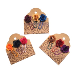 May include: Three sets of decorative paper clips, each featuring a different color of paper rose. The roses are attached to metal clips in various colors, presented on brown tags with a geometric pattern. Colors include orange, black, red, and purple.