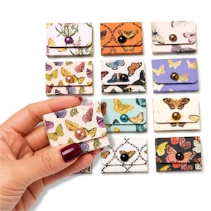 May include: A collection of small, rectangular paper envelopes featuring various butterfly patterns and colors, each secured with a single round, colored bead button closure. The envelopes are arranged in a grid, showcasing diverse prints.