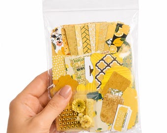 May include: A clear plastic bag filled with a collection of yellow-themed paper ephemera, including die-cut shapes, patterned cardstock strips, small fabric flowers, and a rectangular tag with the printed text KEEP THIS COUPON 302898300.