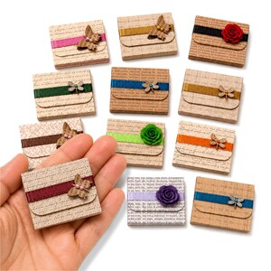May include: A collection of small, rectangular, beige paper boxes featuring a vintage script pattern. Each box is adorned with a horizontal ribbon in various colors, accented by either a small metal dragonfly charm or a fabric flower embellishment.
