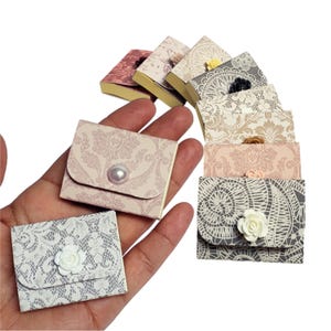 May include: A collection of small, decorative boxes with various floral and lace patterns. Each box has a pearl or flower embellishment. The boxes are arranged in a fan shape and are held in a hand.