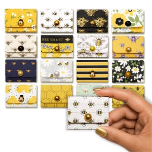 Könnte beinhalten: Eine Sammlung kleiner, rechteckiger Papierumschläge mit verschiedenen Bienen- und Blumenmustern in Gelb, Schwarz, Weiß und Gold. Jeder Umschlag ist mit einem dekorativen Knopf oder einer Perle verschlossen. Ein Umschlag wird zur Ansicht gehalten.