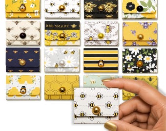 May include: A collection of small, rectangular paper envelopes featuring various bee and floral patterns in yellow, black, white, and gold. Each envelope is secured with a decorative button or bead closure. One envelope is held up to show scale.