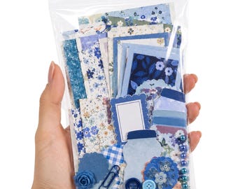 May include: A clear plastic bag containing a collection of blue-themed paper ephemera, including floral patterned cardstock, rectangular frames, scalloped die-cuts, a blue paper rose, a metal paperclip, two round buttons, and a row of small blue beads.