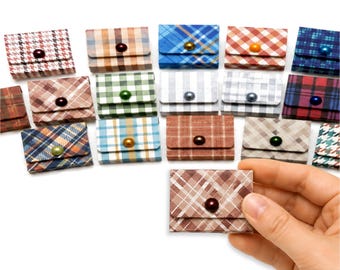 May include: A collection of small, square, envelope-style pouches featuring various plaid and checkered patterns in multiple colors. Each pouch has a single button closure. The text 'Includes One' is visible at the top of the image.
