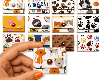 May include: A collection of small, rectangular paper envelopes featuring various cat-themed patterns, including paw prints, colorful yarn balls, and cartoon cats. Each envelope is secured with a single, centered, round, metallic-finish bead button.