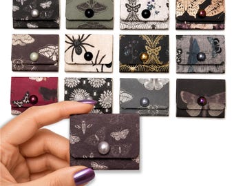 May include: A collection of thirteen small, rectangular paper envelopes featuring various dark, nature-inspired patterns including moths, spiders, and floral motifs. Each envelope is secured with a single, centered, round bead or button fastener.