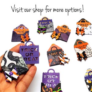 Op de afbeelding: Een verzameling Halloween-thema papieren ambachten, waaronder miniatuur traktatiezakjes en enveloppen. De zakjes zijn paars en oranje, met de woorden "Trick or Treat" uitgesneden. De enveloppen hebben verschillende patronen en strikaccenten. Tekst op de afbeelding luidt "Visit our shop for more options!"
