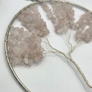 May include: A circular wall hanging with a wire tree design. The tree's branches are decorated with small, pale pink rose quartz stones. The piece is suspended by a silver chain, creating a decorative and natural aesthetic.