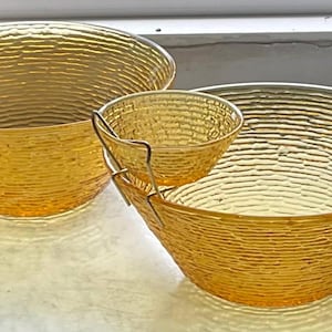 May include: Three amber-colored glass bowls with a textured, ridged design. The set includes two larger bowls and a smaller bowl that hangs from the rim of one of the larger bowls with a metal holder. The bowls are likely for serving.