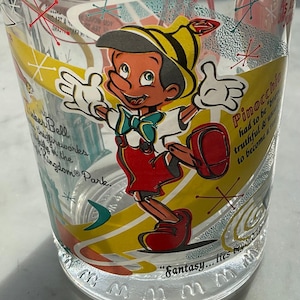 May include: A clear glass cup with a colorful design featuring Pinocchio, a Disney character. The design includes the text "Pinocchio had to be truthful & honest to become a real boy." and "Fantasy... It's beyond your wildest dreams."