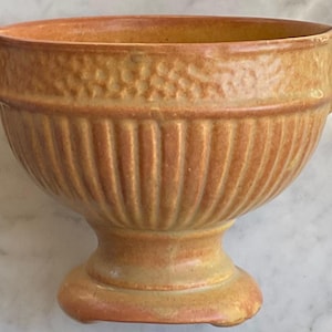 May include: A vintage, peach-colored ceramic bowl with a pedestal base and two small handles. The bowl features vertical ribbed detailing on the lower portion and a textured band near the rim. The overall design is symmetrical and classic.