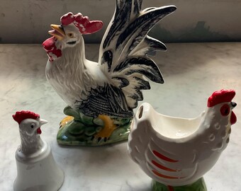 May include: A collection of ceramic rooster and hen figurines. The rooster is white with black and blue tail feathers and a red comb. The hen is white with red accents. A small white bell-shaped figurine with a red comb is also present.