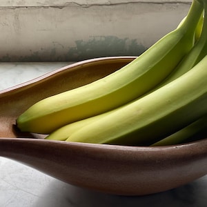 May include: A wooden, boat-shaped bowl filled with a bunch of green bananas. The bananas are still unripe, with a pale green color. The bowl is a warm brown color and sits on a light-colored surface.