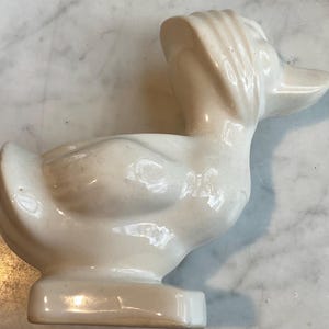 May include: A white ceramic duck planter with a smooth, glossy finish. The duck has a rounded body and a long, curved beak. The planter has a small opening on the top for planting.