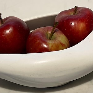 May include: White ceramic fruit bowl with a unique shape and a handle. The bowl is filled with three red apples.