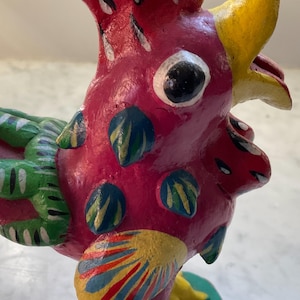 May include: A colorful, hand-painted rooster figurine with a pink body, green and white tail feathers, yellow beak, and blue and yellow accents. The rooster is standing on a green and white base.