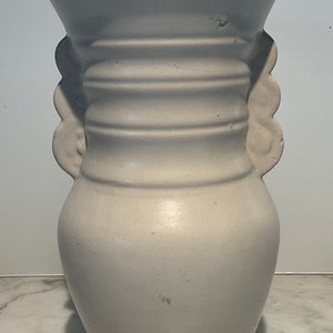 May include: White ceramic vase with a rounded body and a wide mouth. The vase has two handles that are shaped like stylized faces. The vase has a smooth, matte finish.