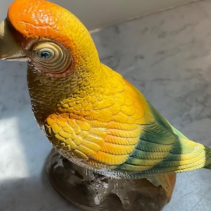 May include: A detailed, hand-painted parrot figurine. The bird features a vibrant color palette of yellow, orange, and green. The parrot has a detailed eye and beak, and is perched on a textured base, showcasing a realistic avian sculpture.