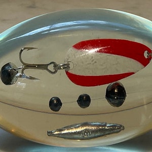May include: A clear plastic egg-shaped paperweight with a red and white fishing lure, a silver hook, and four silver sinkers inside.
