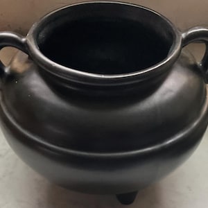 May include: A black ceramic cauldron with two loop handles and three small feet. The pot has a rounded body and a wide opening. The dark color and simple design suggest a rustic or artisanal style, suitable for decorative or functional use.