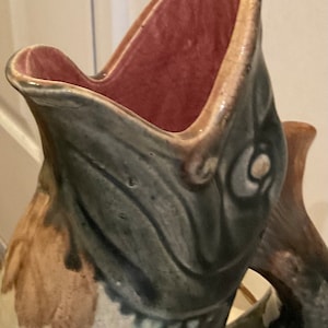 May include: A ceramic pitcher shaped like a fish with a green, brown, and white glaze. The pitcher has a pink interior and a large open mouth.