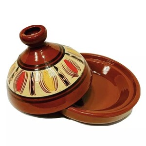 May include: A brown ceramic tagine with a lid. The lid has a white background with a decorative pattern of red, yellow, and black lines and shapes.