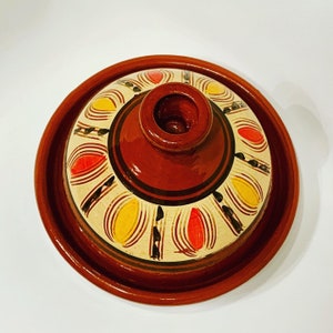 May include: A terracotta tagine lid with a white base and a decorative design of yellow and red ovals. The lid has a brown rim and a small opening in the center.