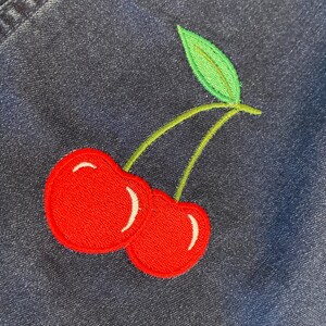 Op de afbeelding: Geborduurde patch met twee rode kersen met witte accenten, groene stelen en een blad. De patch is genaaid op een donkerblauwe denim stof, wat een contrast cre&euml;ert tussen de heldere borduursels en de denim.