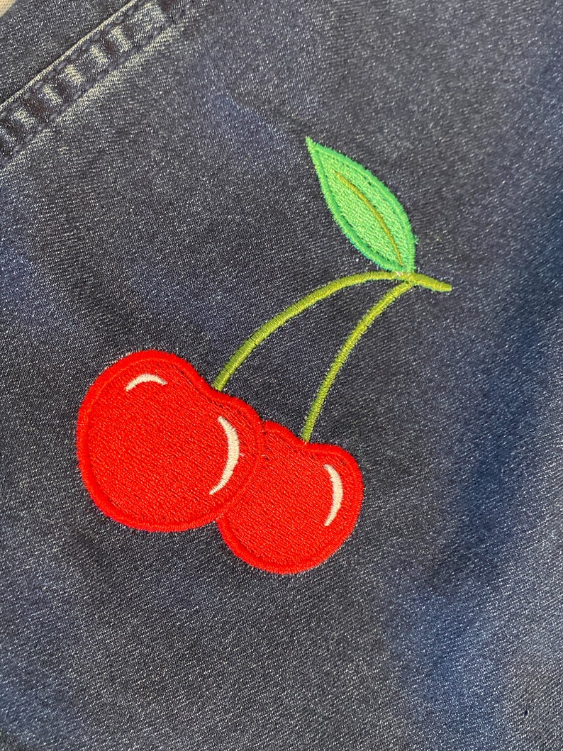 Op de afbeelding: Geborduurde patch met twee rode kersen met witte accenten, groene stelen en een blad. De patch is genaaid op een donkerblauwe denim stof, wat een contrast cre&euml;ert tussen de heldere borduursels en de denim.