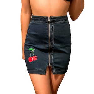 Op de afbeelding: Donkerblauwe denim minirok met een zilveren rits aan de voorkant. De rok heeft een rode kersenpatch met groene bladeren. De rok is een minirok stijl.