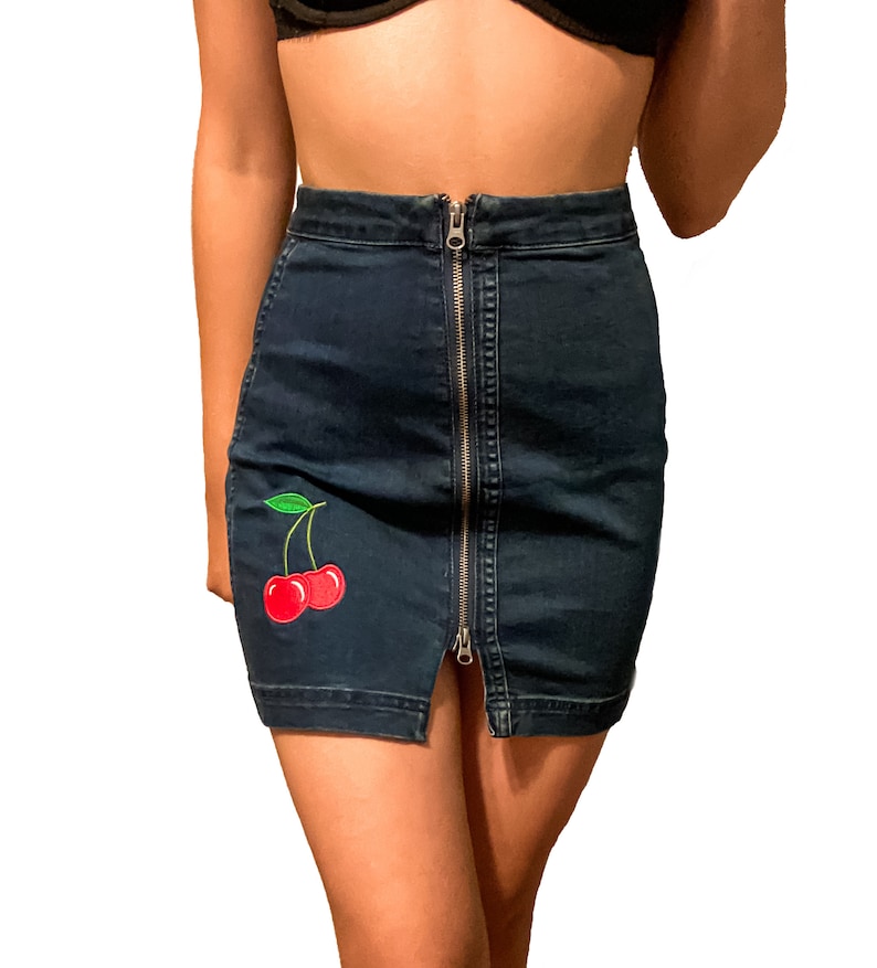 Op de afbeelding: Donkerblauwe denim minirok met een zilveren rits aan de voorkant. De rok heeft een rode kersenpatch met groene bladeren. De rok is een minirok stijl.