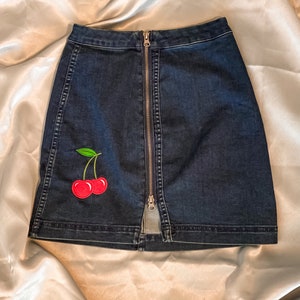 Op de afbeelding: Donkerblauwe denim rok met een ritssluiting aan de voorkant en een kersenpatch. De rok heeft een rood kersendesign met groene bladeren. De rok is een minirok.
