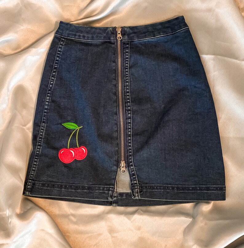 Op de afbeelding: Donkerblauwe denim rok met een ritssluiting aan de voorkant en een kersenpatch. De rok heeft een rood kersendesign met groene bladeren. De rok is een minirok.