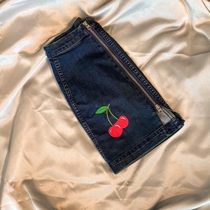 Op de afbeelding: Donkerblauwe denim shorts met een zilveren ritssluiting aan de zijkant. De shorts hebben een rode kersenborduring met groene bladeren. De shorts zijn op een satijnen oppervlak gevouwen.
