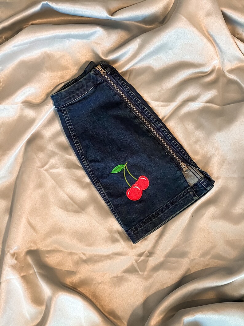 Op de afbeelding: Donkerblauwe denim shorts met een zilveren ritssluiting aan de zijkant. De shorts hebben een rode kersenborduring met groene bladeren. De shorts zijn op een satijnen oppervlak gevouwen.