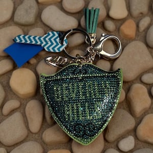 May include: A keychain with a shield-shaped charm covered in green and teal rhinestones. The charm has the words "MOM BOSS" in green. The keychain also includes a blue and white zig-zag key, a teal tassel, and a silver clasp.
