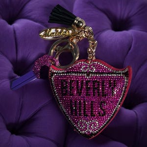May include: A pink and silver rhinestone keychain with the words "BEVERLY HILLS" on a shield-shaped pendant. The keychain has a gold chain, a black tassel, and a purple key.