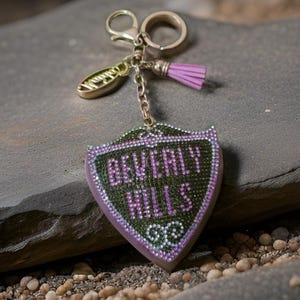 May include: A gold-tone keychain with a pink tassel charm and a green and purple shield-shaped charm that says "Beverly Hills" in rhinestones.