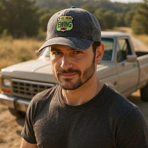 May include: A gray baseball cap with a curved brim. The cap features a patch with the text "EST. 1930 EWING OIL" in white and green. The cap is worn by a person in a gray t-shirt, with a truck in the background.