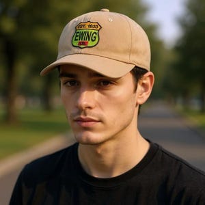 May include: A tan baseball cap with a curved brim. The cap features a patch with the text "EST. 1930 EWING OIL" in green, yellow, and red. The cap is worn by a person wearing a black t-shirt.