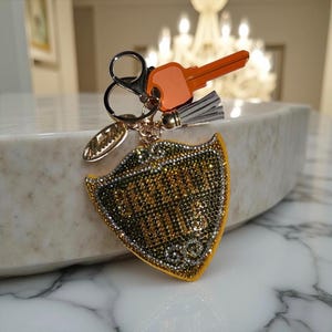 May include: A decorative keychain with an orange key, a shield-shaped charm, and a small tassel. The shield charm is covered in gold and silver rhinestones, with the word "Maria" in gold script. The keychain is attached to a silver clasp.