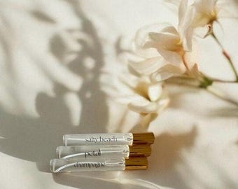 May include: Three clear glass perfume roller bottles with gold caps stacked horizontally. Each bottle features a black label with the text Salty Beach, Petal, or Champagne printed in a serif font. The bottles contain a clear liquid fragrance.