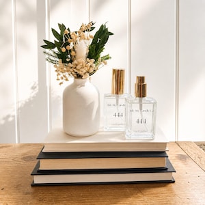 以下が含まれることがあります： 金色のキャップが付いた透明な香水瓶2本。「July Sky Candle Co. 444 eau de parfum」と表示。白い花瓶には、ドライフラワーと緑の小さなブーケが入っています。本の上に配置された静物画。