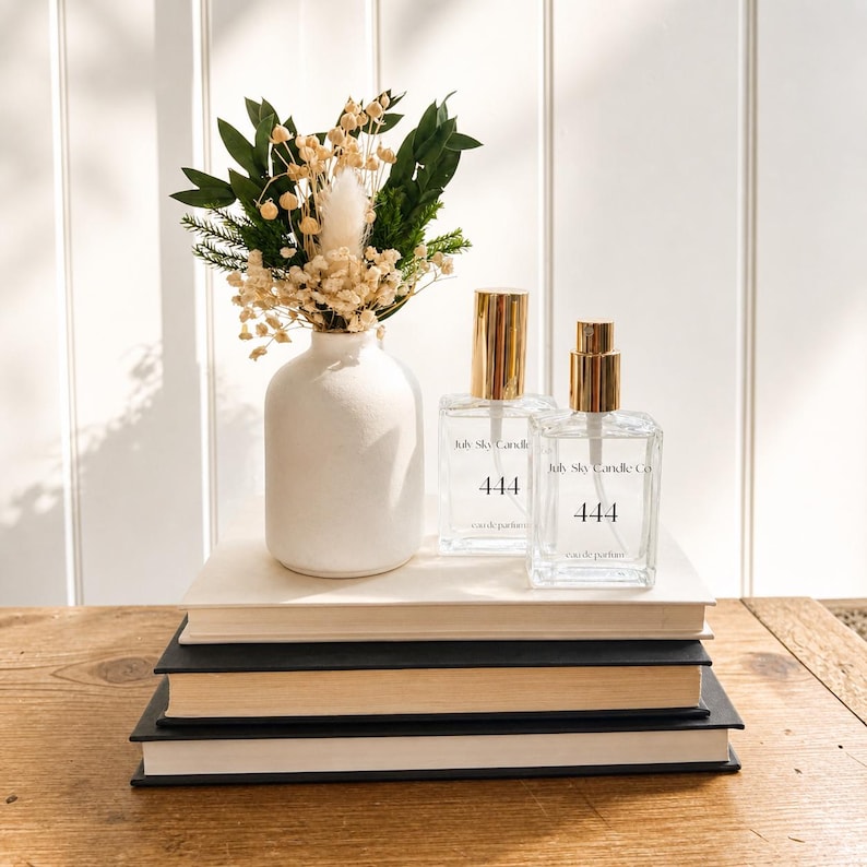 以下が含まれることがあります： 金色のキャップが付いた透明な香水瓶2本。「July Sky Candle Co. 444 eau de parfum」と表示。白い花瓶には、ドライフラワーと緑の小さなブーケが入っています。本の上に配置された静物画。