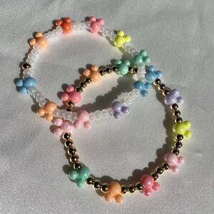 May include: Two beaded bracelets with colorful Mickey Mouse-shaped charms. The charms are in pastel shades of orange, pink, yellow, green, blue, and purple. The bracelets also feature gold-colored beads and white seed beads.