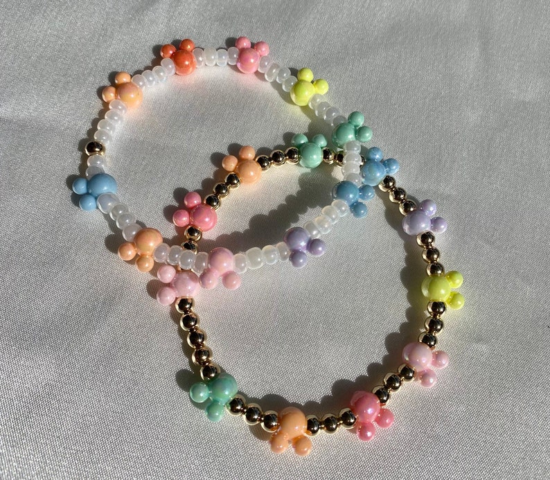 May include: Two beaded bracelets with colorful Mickey Mouse-shaped charms. The charms are in pastel shades of orange, pink, yellow, green, blue, and purple. The bracelets also feature gold-colored beads and white seed beads.