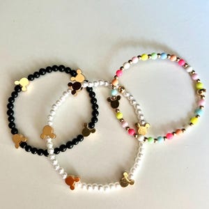 Puede incluir: Tres pulseras con dijes de Mickey Mouse dorados. Una pulsera tiene cuentas negras, otra tiene perlas blancas y otra tiene cuentas de colores.